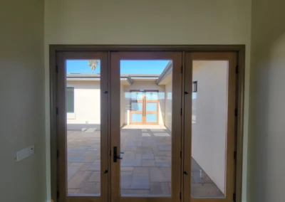A view through wooden glass doors leading to a sunlit patio with a palm tree and modern architecture in the background.