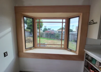 A sunny, wooden bay window with open sides frames a backyard view featuring grass, plants, and a fence.